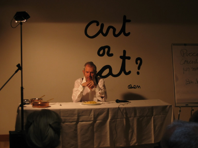 Ben incrocia le mani sopra il piatto di spaghetti. Dietro di lui, sul muro bianco, leggiamo una scritta nera firmata Ben: *Art or Eat*.